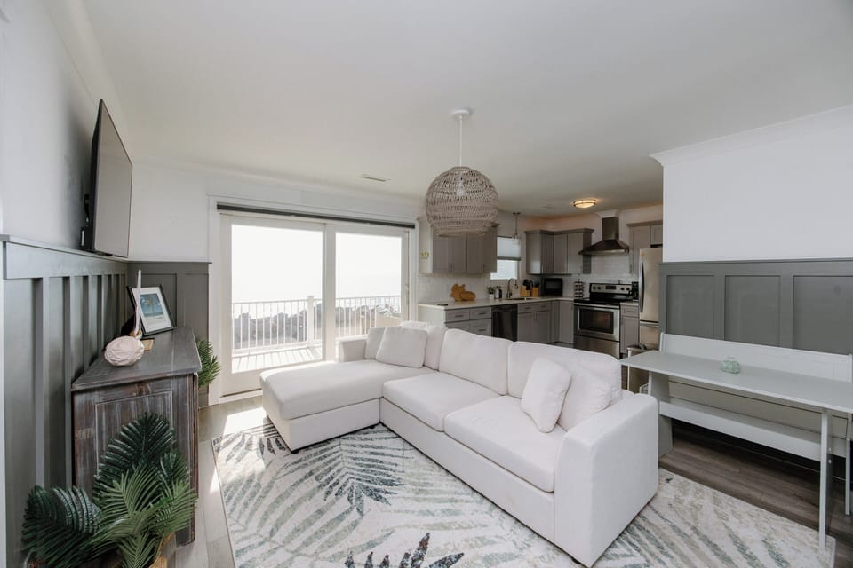 Bright and newly furnished living area featuring large ocean‑view windows, cozy seating, and a Smart TV.