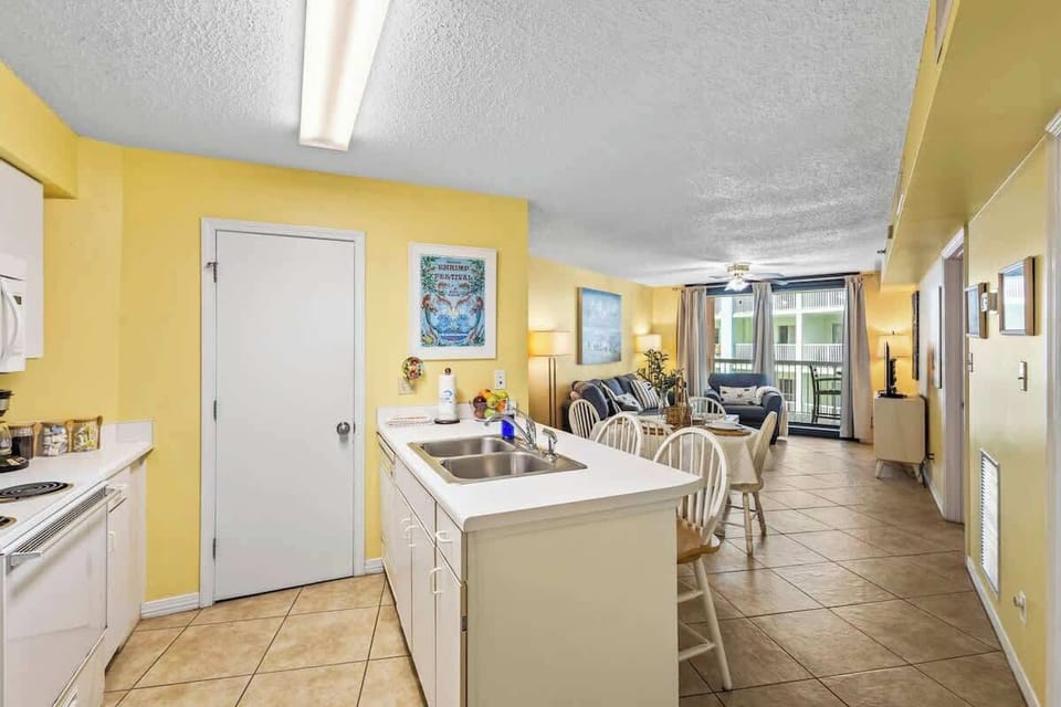 The open kitchen seamlessly flows into the dining and living areas, creating the perfect space for entertaining. A large island with a sink, bright lighting, and beachy décor make it a welcoming spot for meals and gatherings.