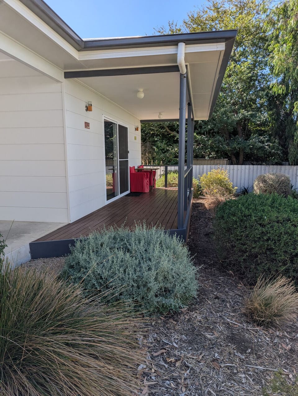 A cozy outdoor area of a house with a wooden deck. There is a red seating set on the deck, and the surrounding area features various green shrubs and plants. The house has light-colored siding and a covered porch, with trees and a fence visible in the bac