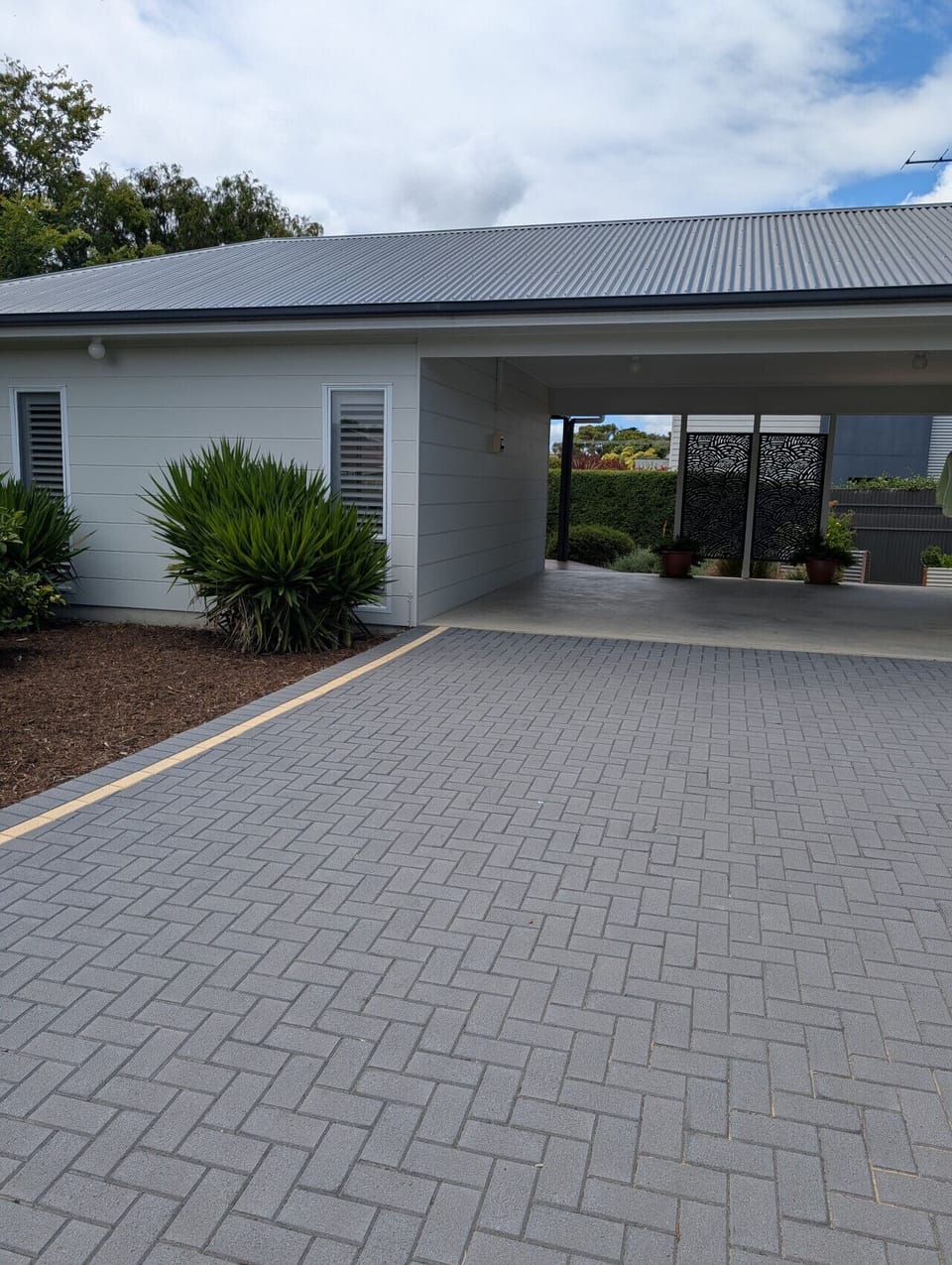 A modern house with a grey corrugated roof and light-colored walls. There is a paved area in the foreground with a neat herringbone pattern, and some greenery including plants near the house, adding a touch of nature to the scene. The overall setting appe