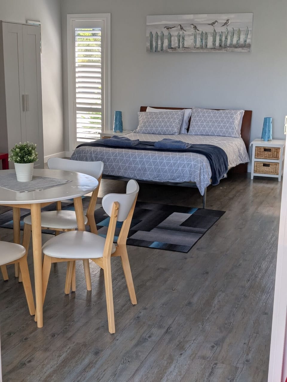 A cozy studio room with a bed adorned with patterned bedding and pillows, a small table with white chairs, a white cabinet, a window with shutters, and a decorative piece of art on the wall. The room also features a small plant on the table and some blue 