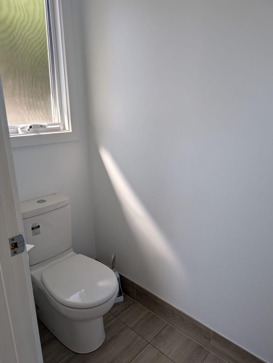 A small, clean bathroom with a white toilet. The room has white walls and a window with a frosted glass that allows some light to enter, casting a shadow on the wall. The floor is tiled with a light brown wood-like pattern.