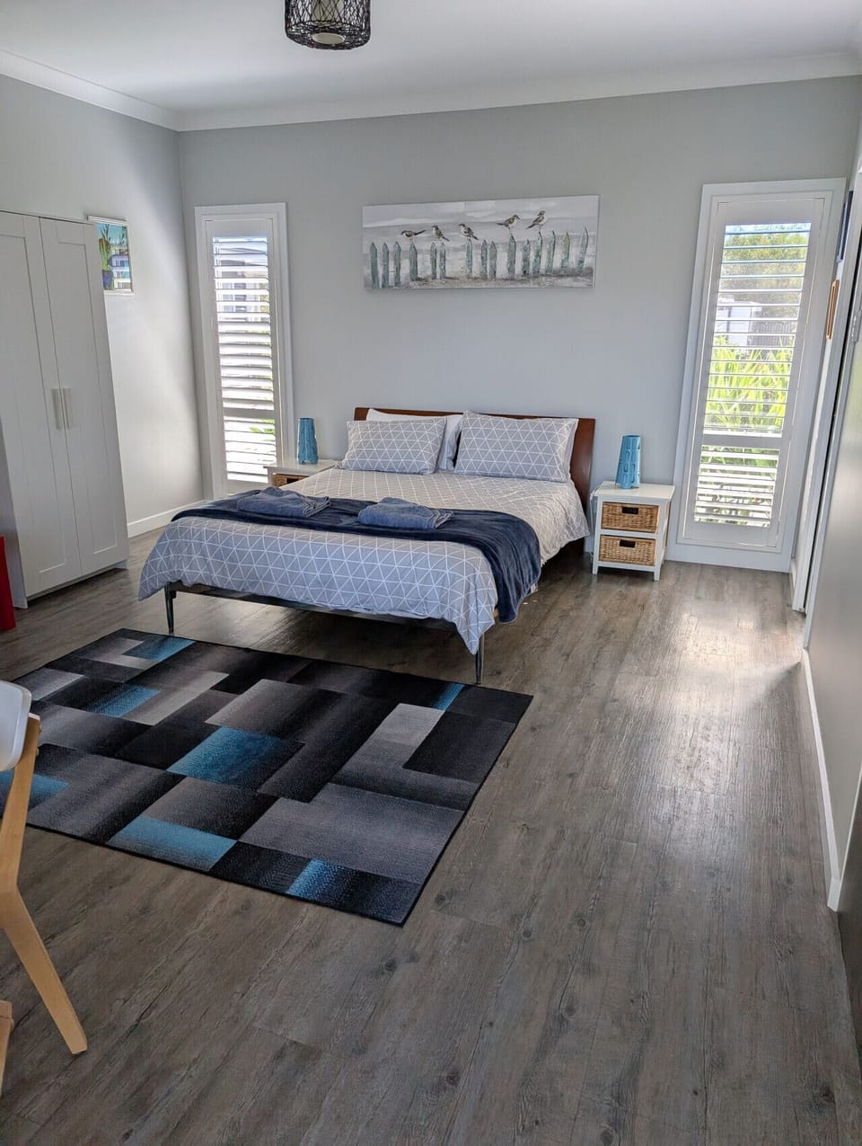 A neatly arranged bedroom with a bed covered in patterned bedding and a blue throw blanket. The room features light-colored walls, wooden flooring, and two windows with shutters that let in natural light. There is a decorative rug on the floor, a white ca
