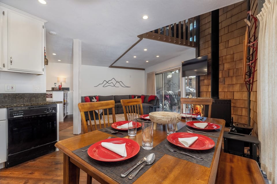 Gather with friends and family in this beautifully appointed condominium, where a warm wood-burning stove, inviting sectional sofa, and mountain-inspired décor create the perfect après-ski retreat. The open-concept kitchen and dining area, set with vibrant red dishware and bathed in natural light from sliding glass doors, offers a seamless blend of comfort and style for your mountain getaway.