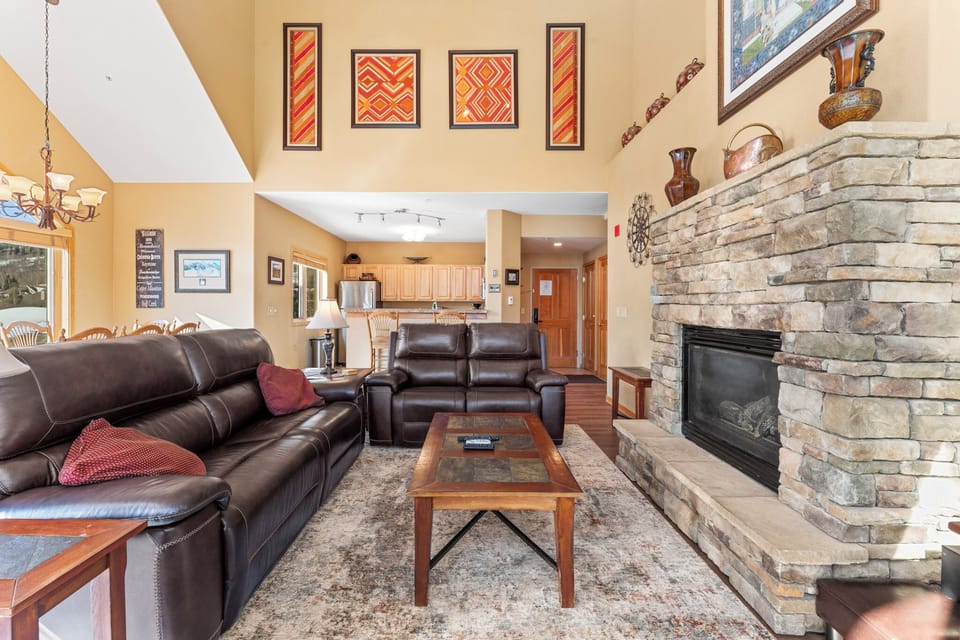 Spacious living area with vaulted ceilings, large windows providing ample sunlight, and electric reclining sofas surrounding the cozy fireplace.