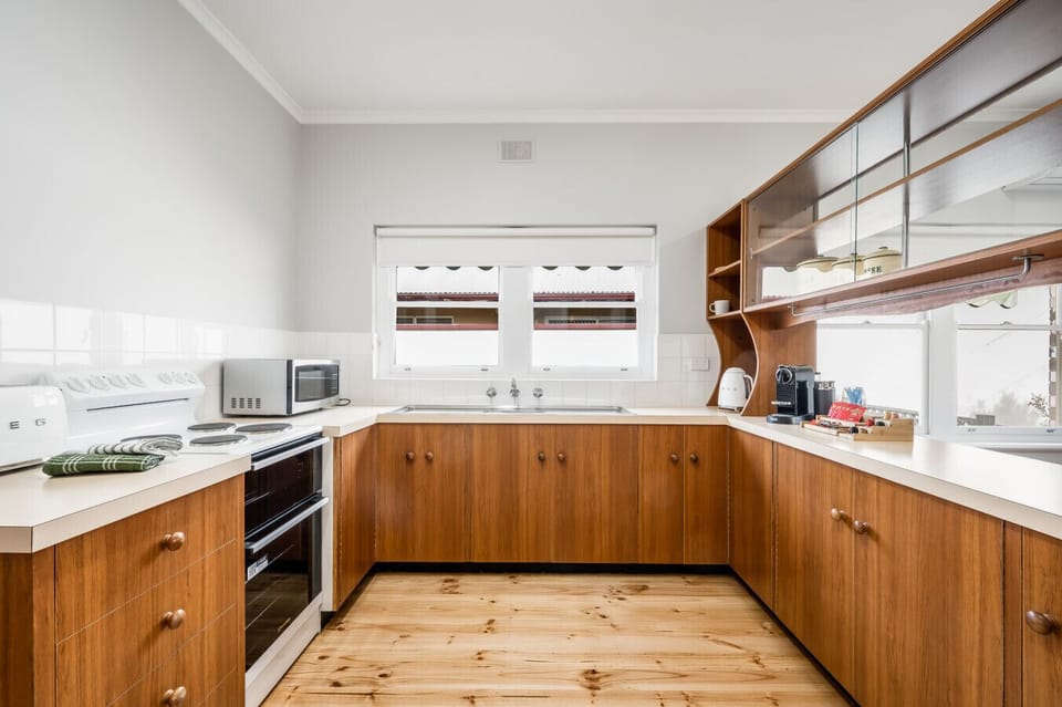 A clean and well-organized kitchen with wooden cabinets and a light-colored countertop. The kitchen features a stove, a microwave, and various kitchen appliances. The wooden flooring adds a warm, rustic touch to the space, and there are windows that allow
