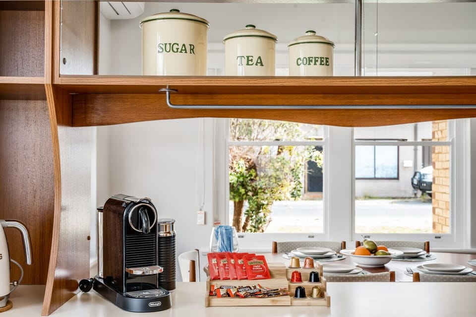 On a wooden shelf, there are three labeled canisters for sugar, tea, and coffee. Below the shelf, there is a De`Longhi coffee machine on a countertop, along with a variety of coffee pods displayed in a holder. In the background, there is a dining table se
