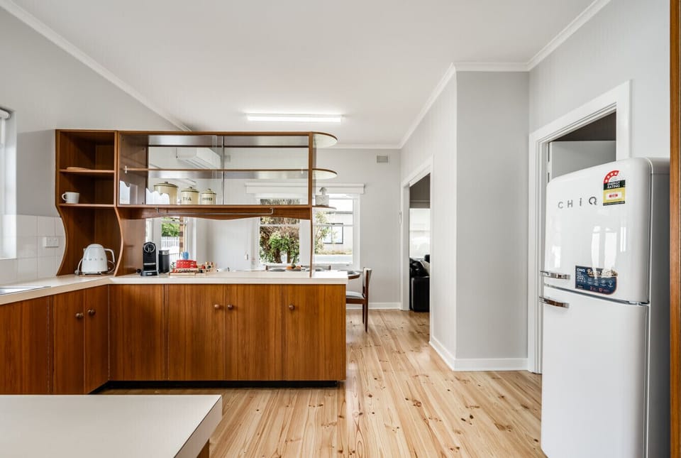 A stylish kitchen with wooden cabinetry and a light wood floor. The kitchen features a unique, retro-style refrigerator labeled `CHiQ` and modern appliances like a kettle and coffee maker. The space has a clean, minimalist design with light-colored walls,