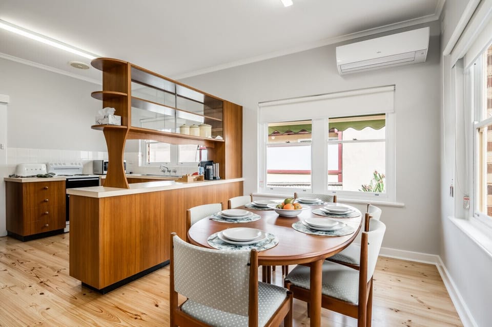 A stylish dining area adjacent to a kitchen with wooden cabinetry and a unique, mid-century modern design. The dining table is set with plates and a bowl of fruit, and the room features light-colored walls, a wooden floor, and large windows that let in na