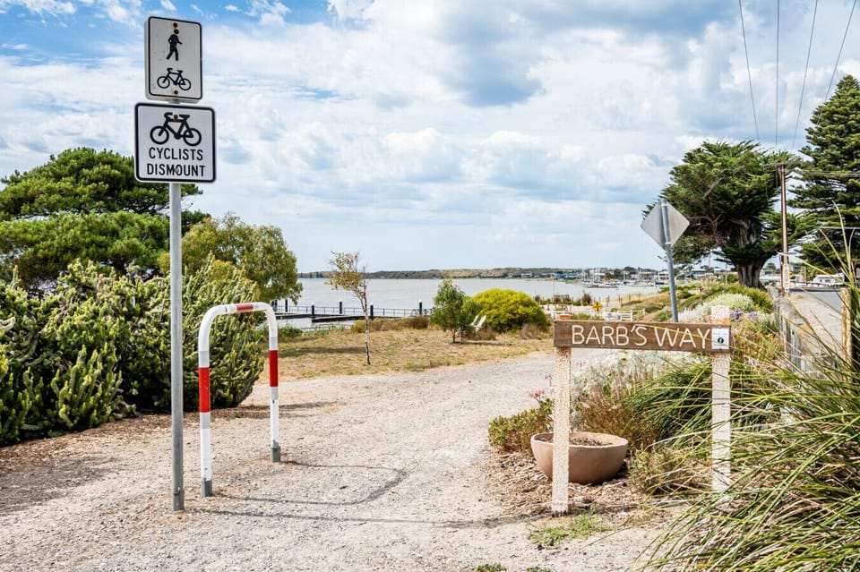 A pathway with a `Cyclists Dismount` sign and a pedestrian sign, along with a barrier. There is also a sign that reads `Barb`s Way,` indicating a named path. The scene includes some greenery and a view of a body of water in the background under a partly c
