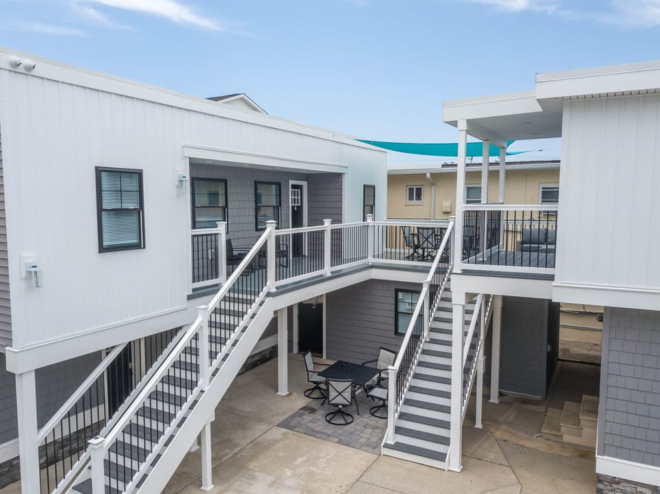 Modern beach-style condo with spacious shared patio, outdoor seating, and shaded upper decks - perfect for relaxing after a day by the shore. Clean, coastal design and easy access via dual staircases.