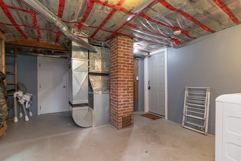 Basement area with exposed ductwork, brick pillar, concrete floor, and laundry appliances near a white door.