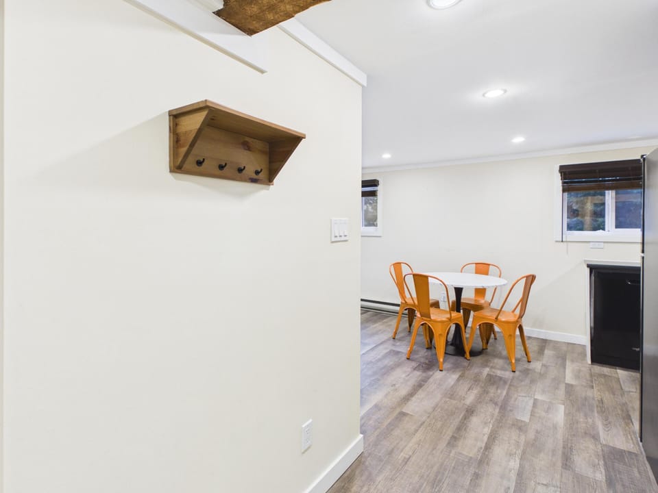 Wall-mounted wooden shelf with hooks hangs on creamy white wall above wood-look vinyl flooring near the dining area.