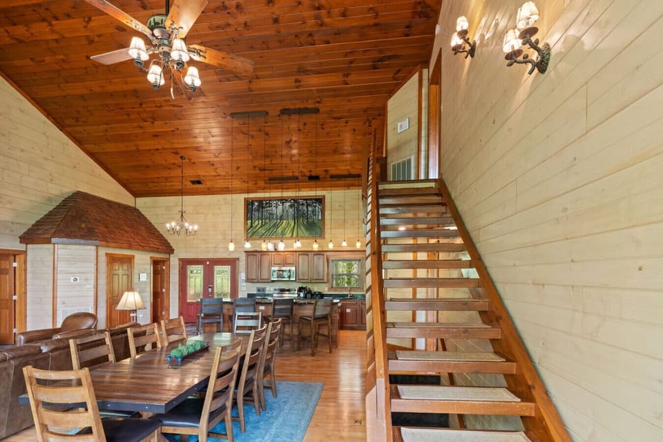 This angle highlights the spacious dining area and striking wooden staircase that leads to the loft and upper bedroom, all set beneath a vaulted cedar ceiling.