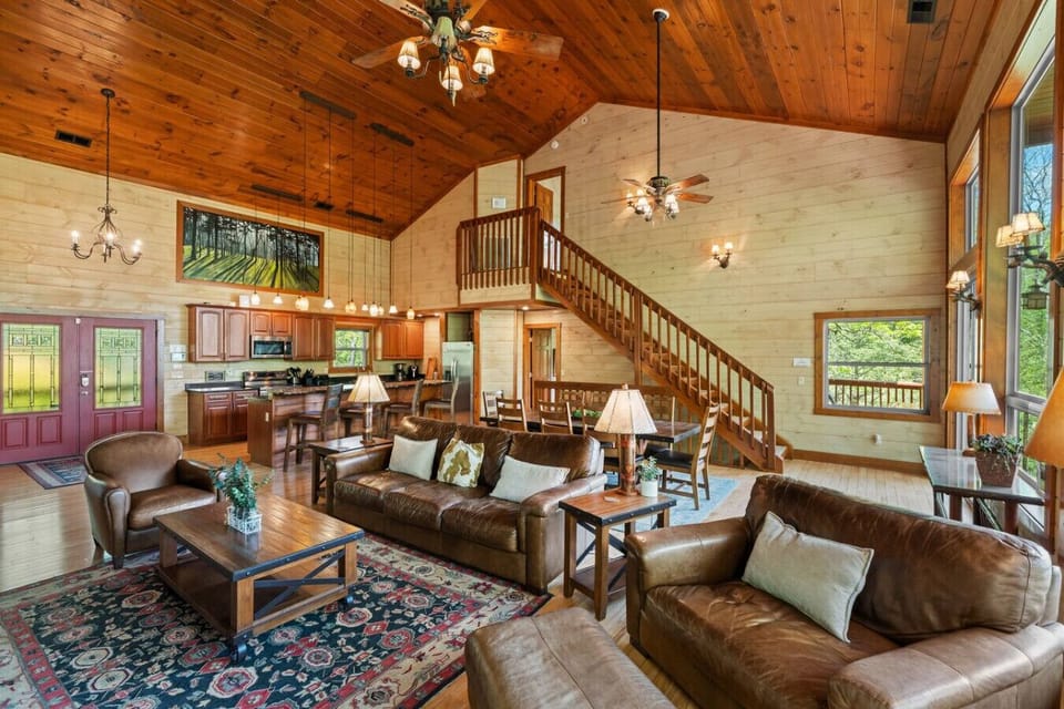 Gather in the open-concept living room featuring a soaring wood-paneled ceiling, plush leather seating, and expansive views of the forest through floor-to-ceiling windows.
