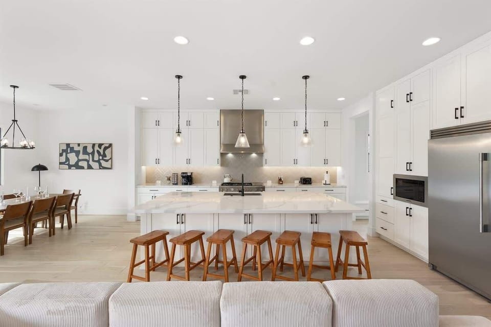 Airy kitchen anchored by a generous island with multiple stools, pendant lighting above, and stainless appliances. The open layout invites cooking, gathering, and lingering conversations while meals come together.