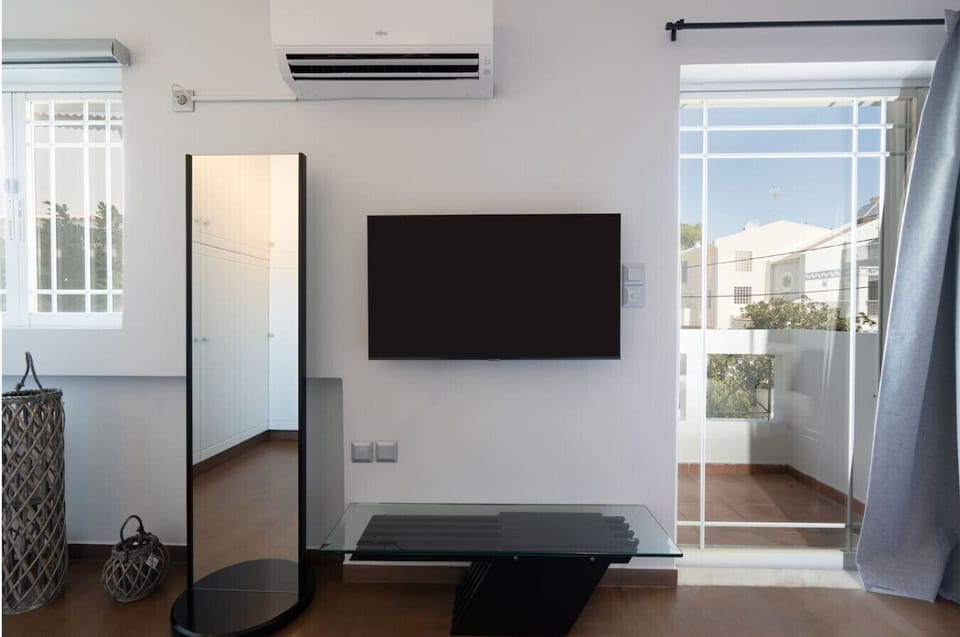 A final bedroom angle — smart TV, full-length mirror and bright Glyfada light streaming through the window. Every room, photo-ready. Booking.com tag: Bedroom