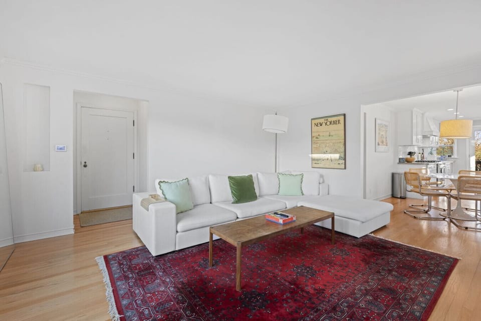 A large white sectional sofa with green accent pillows provides plenty of seating, complemented by a wooden coffee table over a vibrant red Persian-style rug.