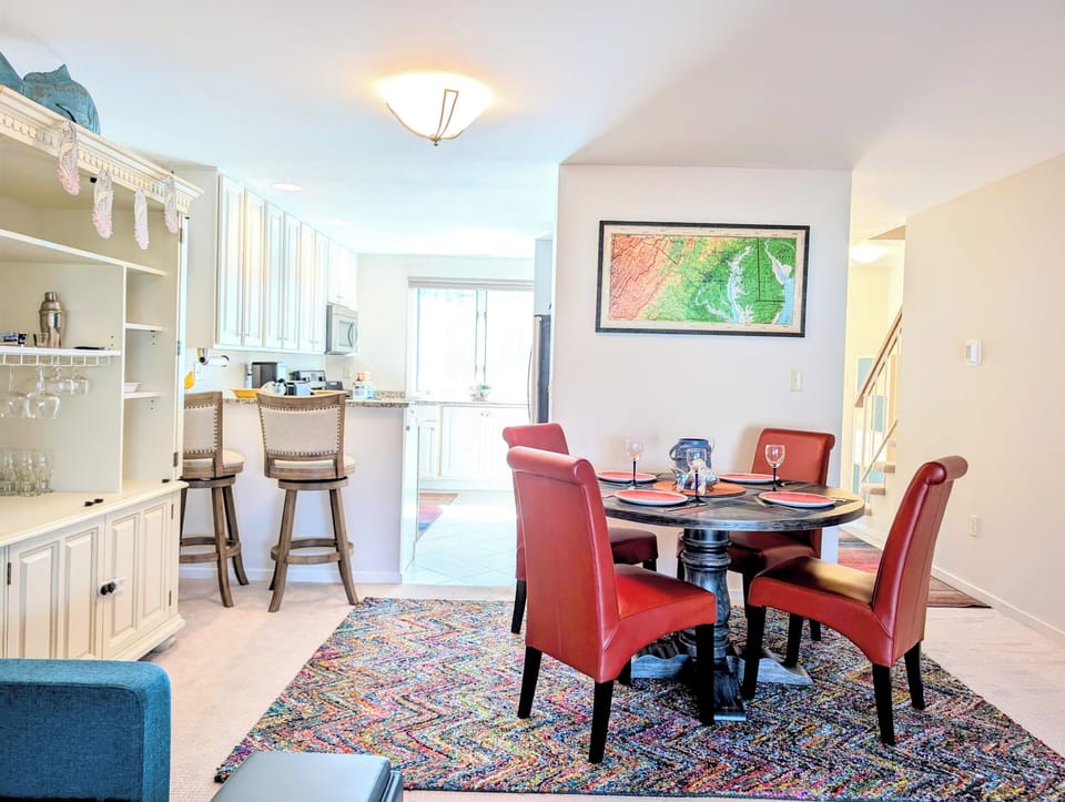 719A Treetop Lane - Living and Dining Connection - Wide view of a dining room with red chairs on an ornate patterned rug, adjacent to a white kitchen and a large cream-colored bookshelf hutch in a bright townhome interior.