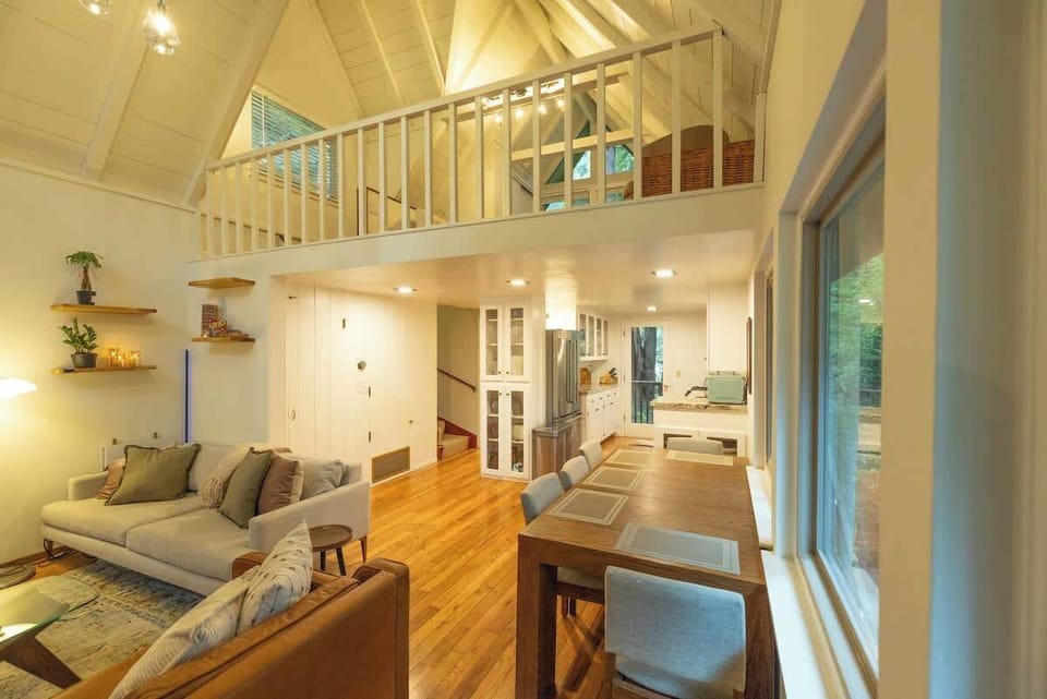 This bright open-concept living area features soaring ceilings, a charming loft above, cozy seating, and a spacious dining and kitchen area—creating an inviting space to gather and relax