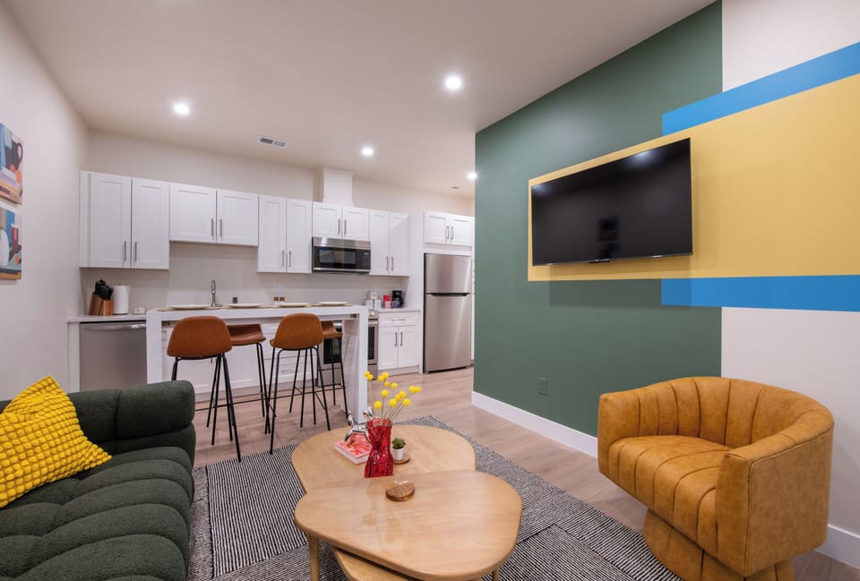 - Bright open concept living space with smart TV, velvet sofa, and modern mustard accent chair
- Kitchen boasts stainless appliances, bar seating, and crisp white cabinetry
- Warm wood floors and bold color blocks create a vibrant artistic vibe