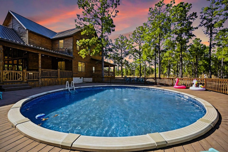 Escape to serenity in our luxury Broken Bow cabin, featuring a sparkling pool amidst towering pines, under a sunset sky. Perfect for a tranquil retreat!