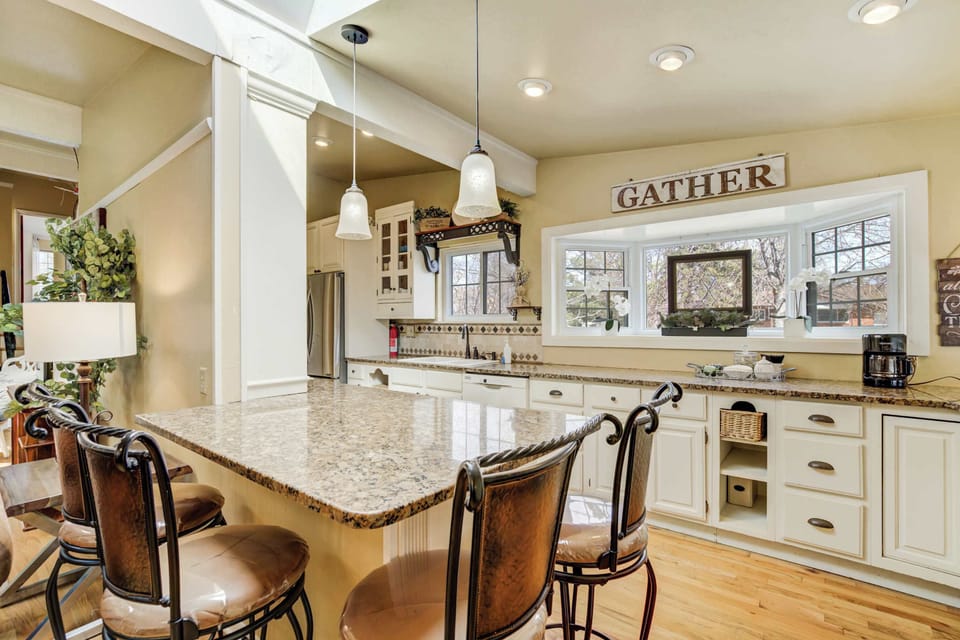 Pull up to the stone-topped island under pendant lights—ideal for quick breakfasts, snacks, or plating dinner together. White cabinetry and the “GATHER” sign set a welcoming mood.