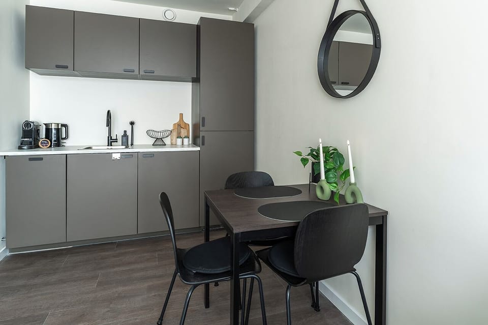 Modern kitchenette with sleek grey cabinets, sink, coffee machine, kettle, and a cozy dining table for two in a clean, stylish space.