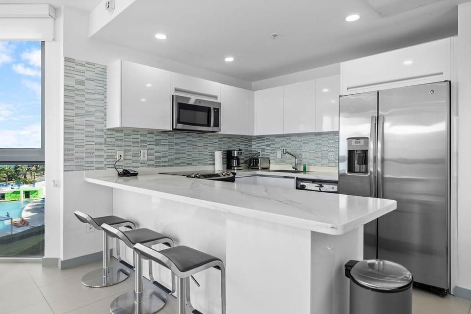Your modern kitchen features sleek white cabinetry, gleaming countertops, and stainless appliances—perfect for whipping up your favorite meals while soaking in tropical views through the window.