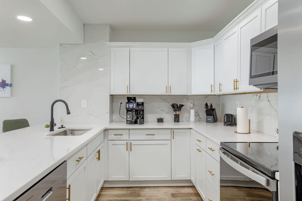 This modern kitchen features white cabinets, sleek countertops, and stainless steel appliances. A coffee maker and various kitchen utensils are provided for your convenience.