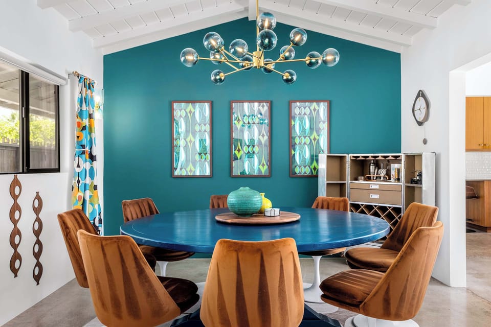 Sunlit mid-century dining room - round blue tulip table for eight, brass Sputnik chandelier, teal accent wall, and built-in bar cart. Dinner here feels like an event.