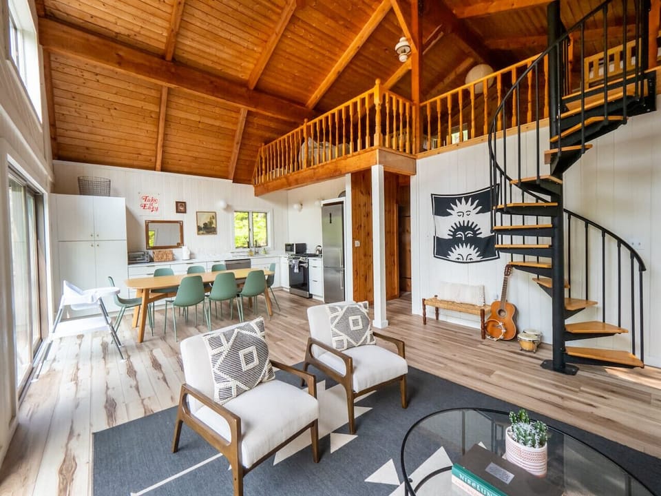 Bright open-concept living room with vaulted wood ceilings, spiral staircase to loft, two accent armchairs, and a view toward the dining area for 8 and kitchen.