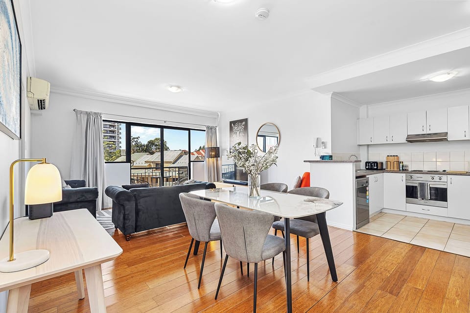 Enjoy an open living and dining area with polished wood floors, modern gray chairs, and a sleek white kitchen in the background.