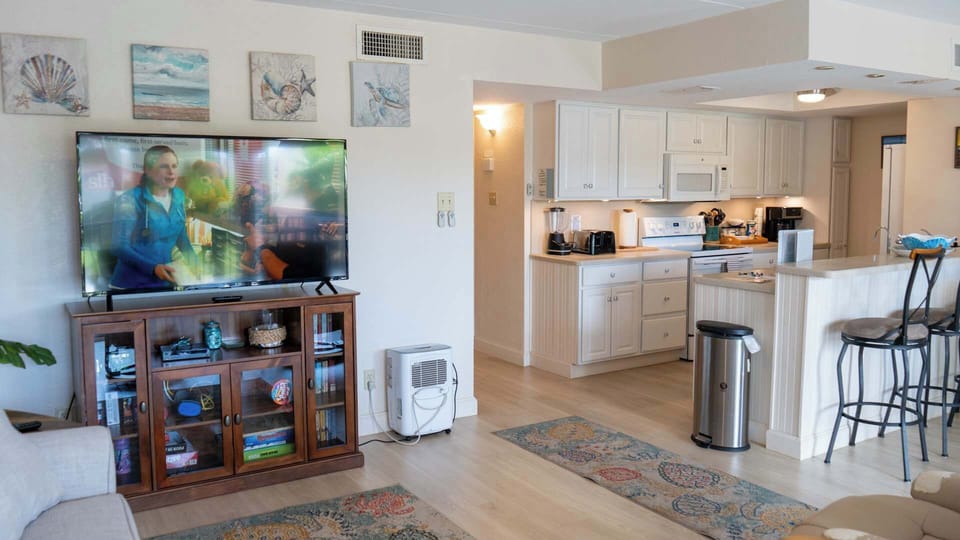 Relax in the open living area featuring a large flat-screen TV, cozy seating, and coastal décor. The space flows seamlessly into a fully stocked kitchen with bar seating, perfect for meals, entertaining, or unwinding after a day at the beach.