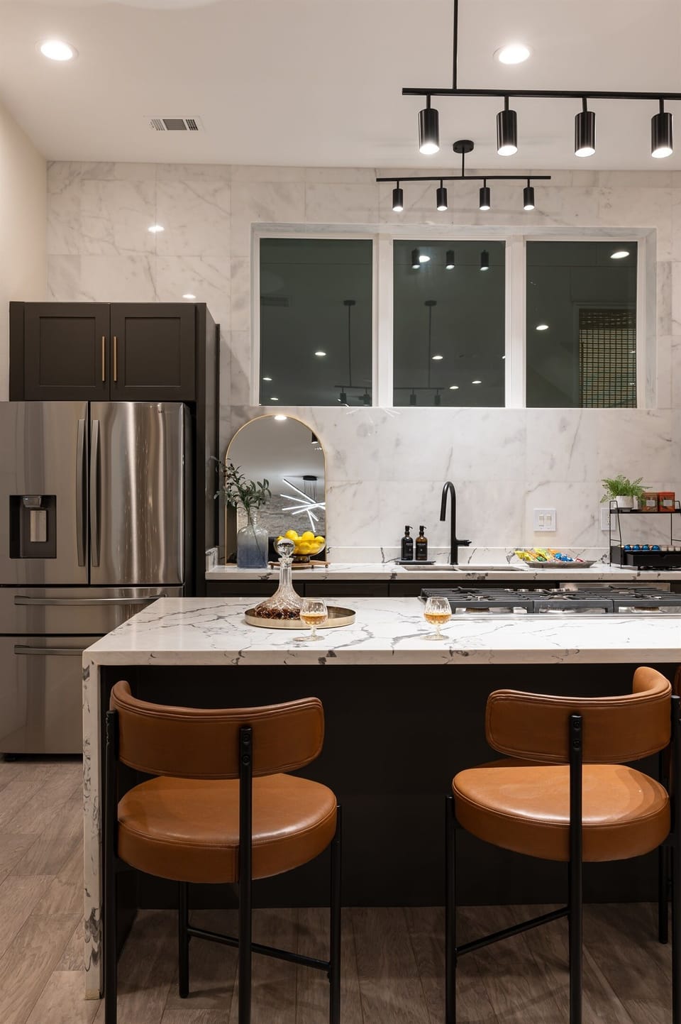 A kitchen with a large island, a stainless steel refrigerator, and dark cabinets. The kitchen has a large window with a view of the city.