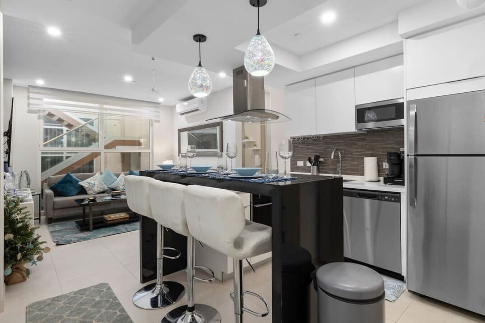 A modern, open-concept living room and kitchen. The kitchen has a black island with white barstools and a white countertop with a stovetop. The living area has a gray sofa with blue pillows and a large window.