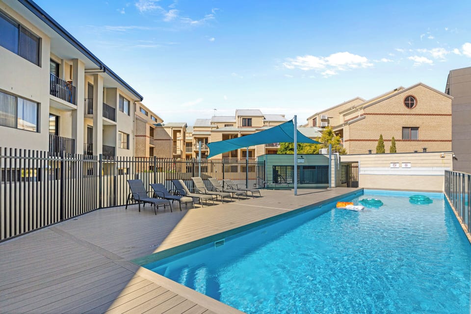 Enjoy the sparkling outdoor pool surrounded by modern apartment buildings on a sunny day, complete with lounge chairs for relaxation.