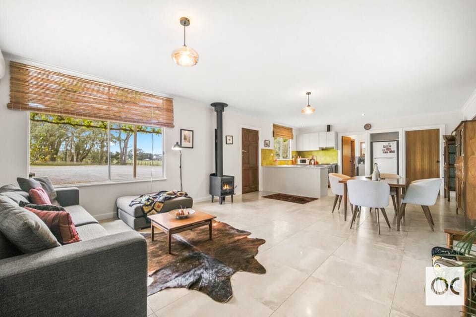 A modern, open-plan living space with a cozy and stylish interior. It features a comfortable gray sofa with colorful cushions, a wooden coffee table on a cowhide rug, and a wood-burning stove for warmth. The area also includes a dining space with light-co
