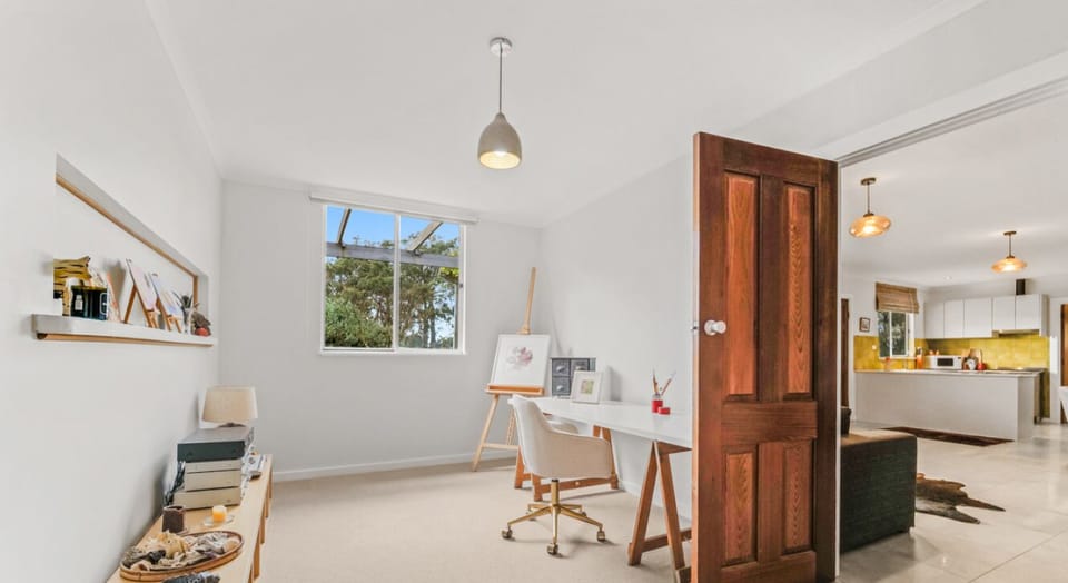 A bright and airy room with a large window allowing natural light to flood in. The room features a wooden door, a desk with a chair, and a shelf displaying various items. Through the open door, another room with a modern kitchen area is visible, adding to