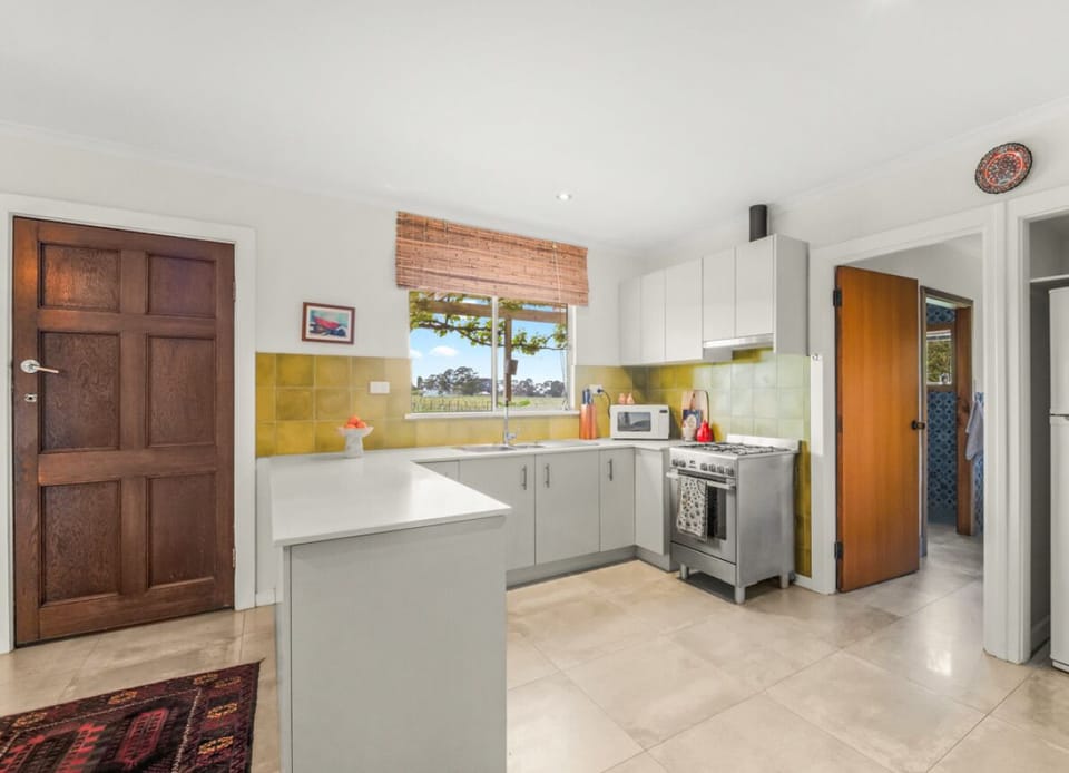 A cozy kitchen with a mix of modern and rustic elements. It features light-colored cabinets, a stainless steel stove, a microwave on the counter, and a window with a bamboo blind offering a view of the outdoors. The kitchen has a wooden door, a patterned 