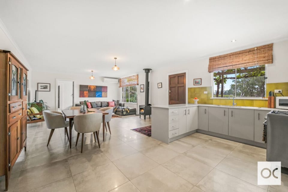 A spacious, open-plan living area with a kitchen featuring light gray cabinets and a yellow backsplash. There is a dining table with chairs, a cozy seating area with a colorful piece of wall art, and a wood stove for warmth. The space has large windows wi
