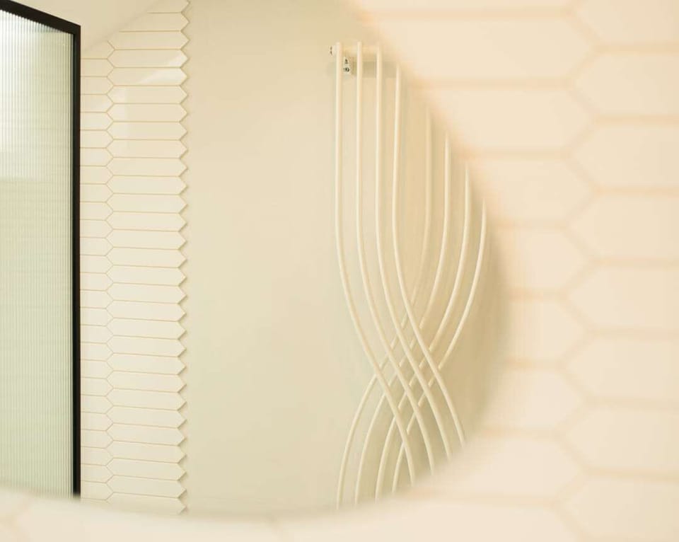 Artful bathroom reflection showcasing a sculptural white towel radiator, soft tile patterns, and a frosted glass shower screen — clean lines meet calm tones.