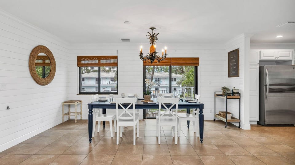 Bright and coastal dining area with a table set for eight, offering views of the charming homes and condos along the street. Features include dinner service for eight, a bar cart ready for cocktail hour, and access to the kitchen for seamless entertainment