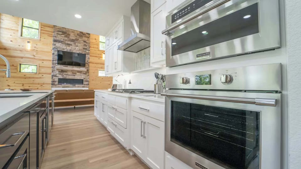 Sleek modern kitchen with stainless steel appliances, bright white cabinetry, and a cozy open layout leading into a wood-accented living area with a stone fireplace.