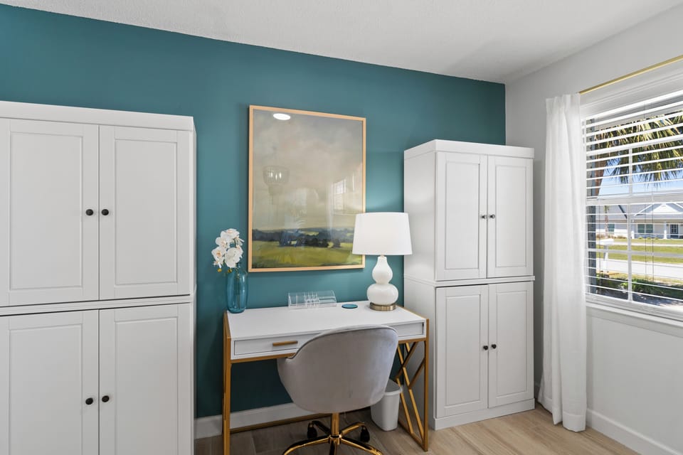 Your stylish workspace awaits with a cozy teal accent wall, modern white furniture, and abundant natural light streaming through the window.