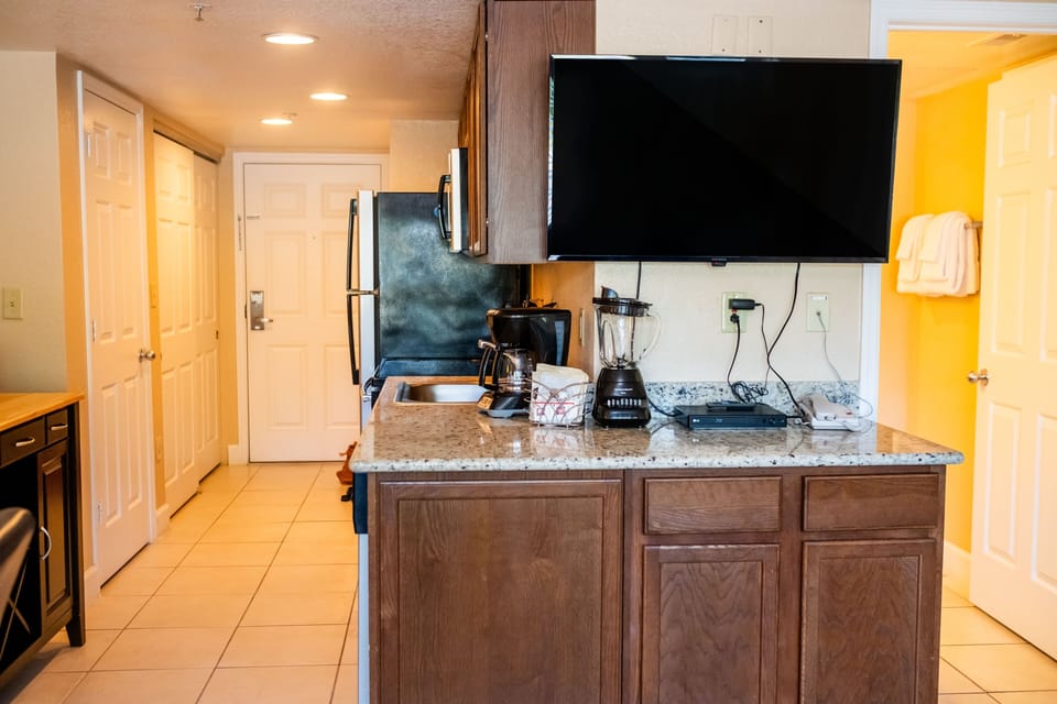 This modern kitchen features a spacious layout with stainless steel appliances, a coffee maker, and a large TV mounted above the granite countertop, perfect for watching while preparing meals.