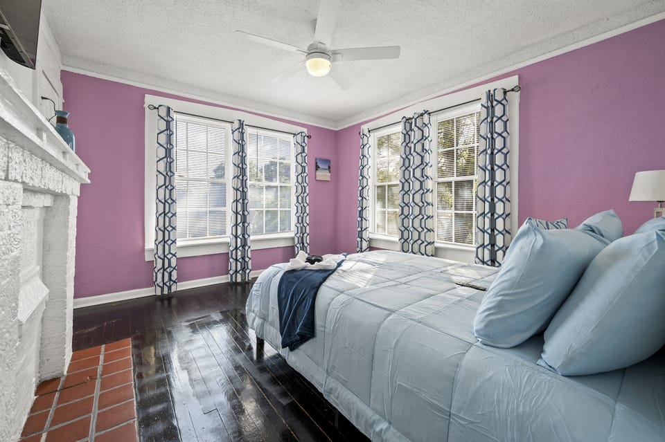 This charming bedroom boasts soothing light purple walls complemented by rich dark wooden floors. The inviting bed is adorned with a plush light blue comforter and matching pillows, creating a serene sleeping environment. 