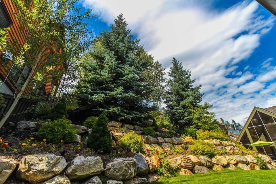 Lush landscaping with vibrant flowers and sturdy trees enhances the beautiful outdoor setting, ideal for summer BBQs or hot tub access.