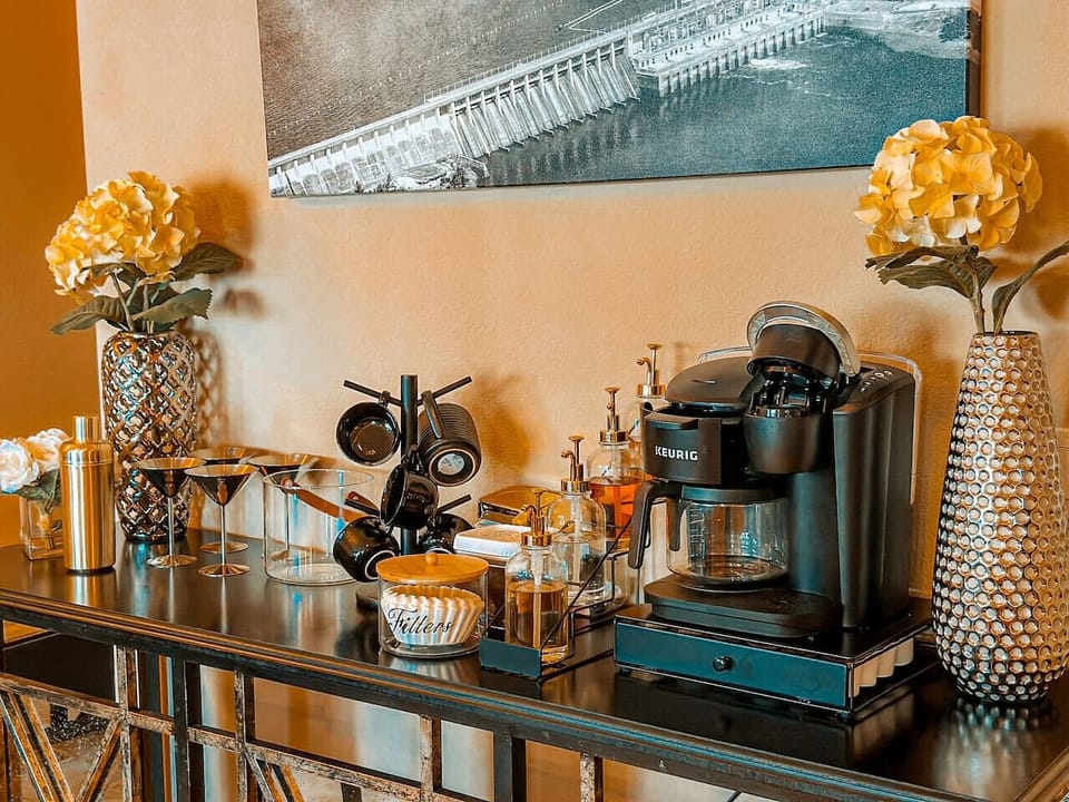 This inviting coffee bar offers a fully equipped station for your morning brew, featuring a Keurig coffee maker and an array of accessories. Complementing the setup, two elegant flower arrangements add a touch of freshness and charm to the space.