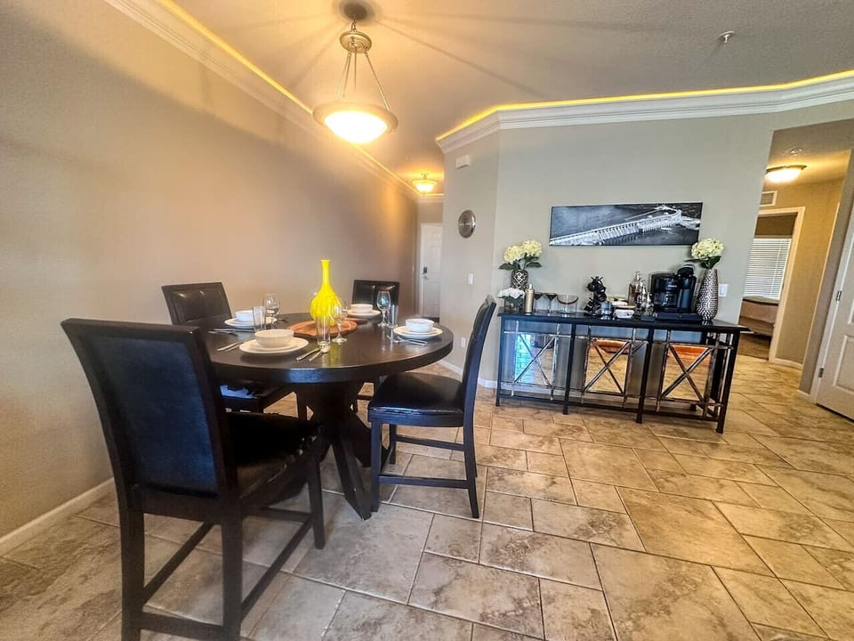 This elegant dining area features a round table set for a welcoming meal, complemented by sophisticated lighting and stylish decor. Adjacent to the dining space, a well-appointed bar area offers a coffee maker and drinkware for your convenience and e